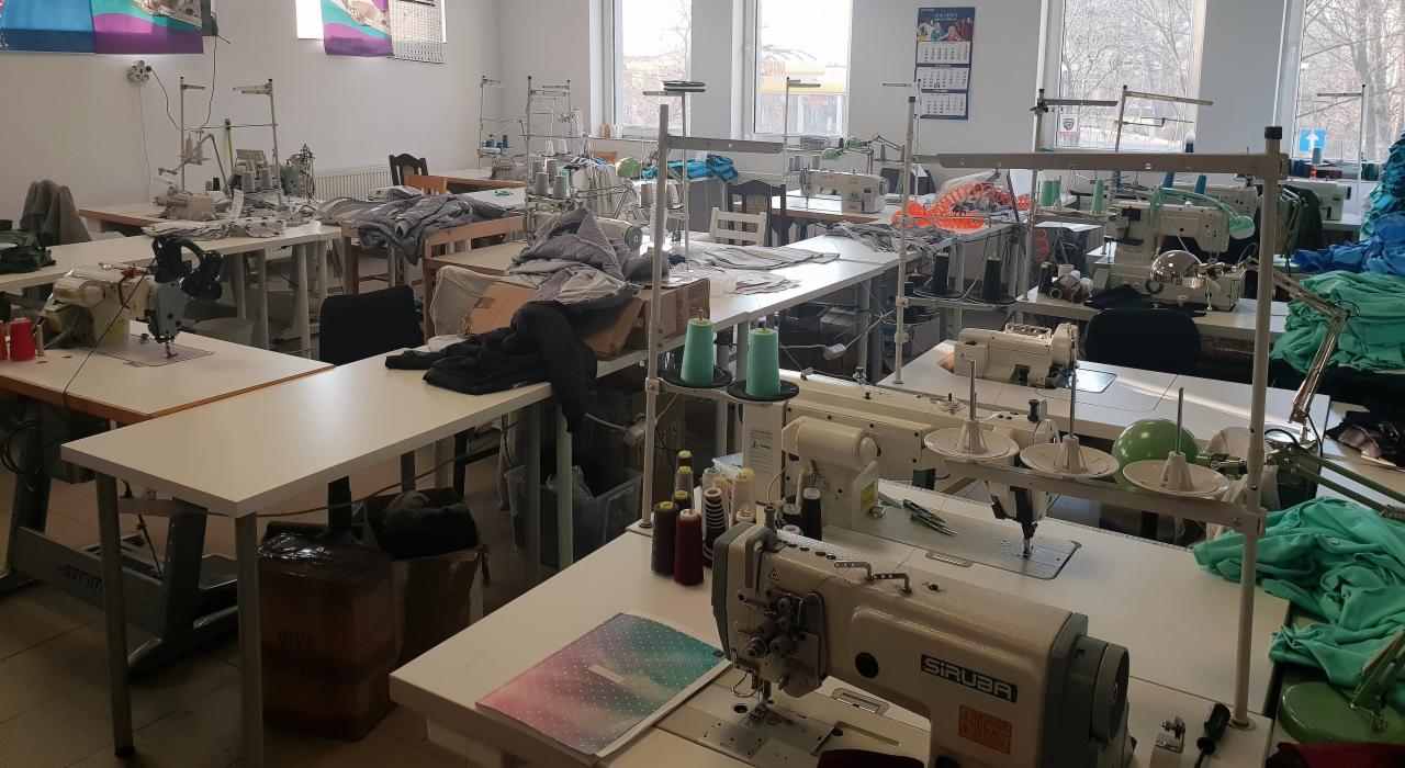 Szwalnia: Wnętrze pracowni krawieckiej z wieloma stanowiskami pracy wyposażonymi w maszyny do szycia, nici i materiały, rozłożone na białych stołach.