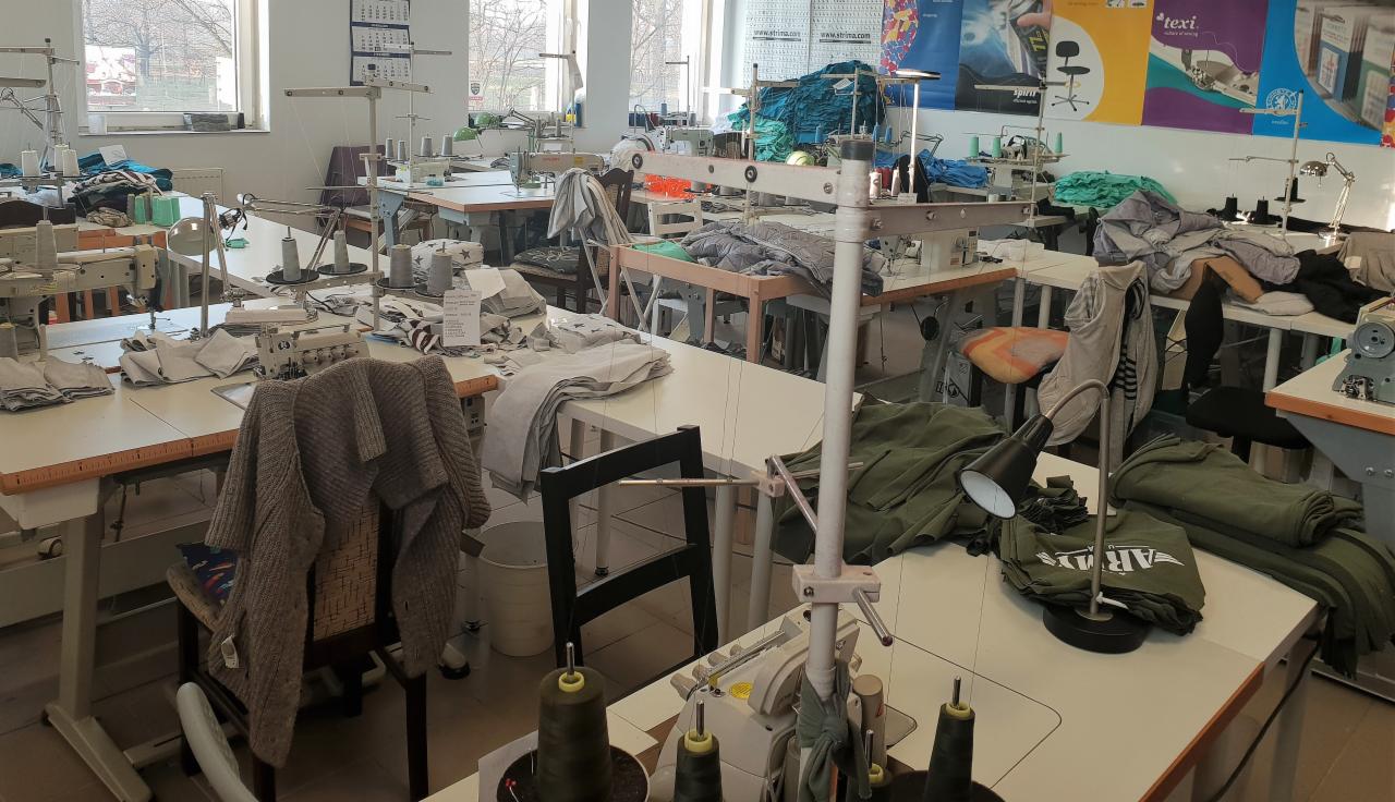 Szewska pracownia: liczne maszyny do szycia na stołach, stosy materiałów i ubrań w różnych kolorach, widoczne szpule nici i lampki biurowe, kalendarz na ścianie, okno w tle.