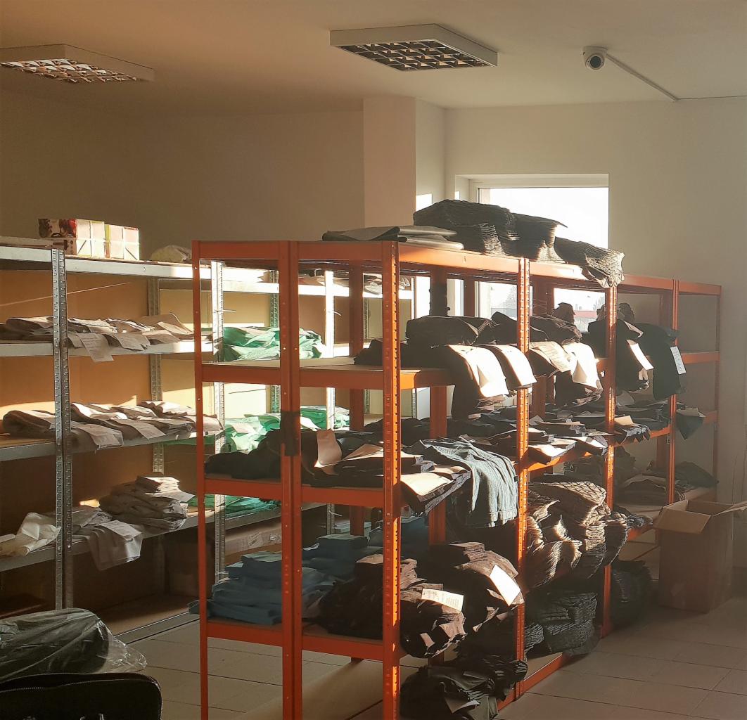 Magazyn szwalni w Legnicy: pomarańczowe regały z poukładanymi stosami materiałów i wykrojów odzieży dziecięcej w różnych kolorach, oświetlone światłem dziennym.