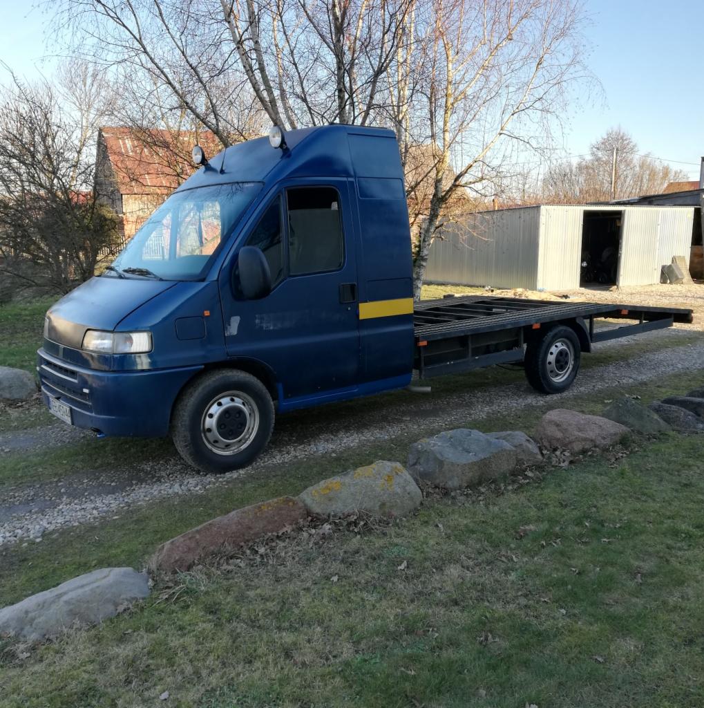 Niebieski samochód dostawczy z platformą lawety, zaparkowany na żwirowej drodze przed budynkiem gospodarczym i domem w tle.