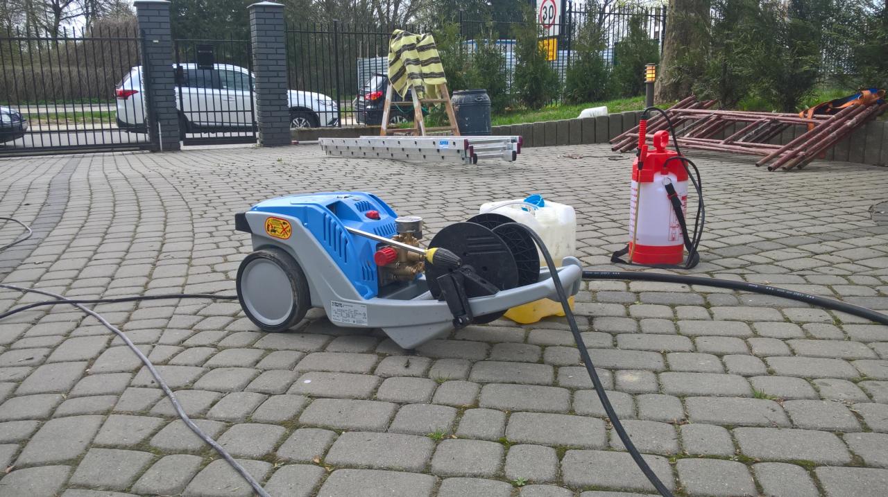 Profesjonalna myjka ciśnieniowa na kostce brukowej, z widocznymi akcesoriami i drabiną w tle, sugerująca przygotowania do czyszczenia elewacji.