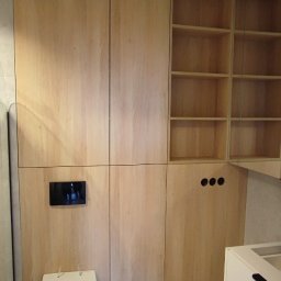 M&R MEBLE - Nowoczesna łazienka z zabudową z jasnego drewna, ukrywającą spłuczkę i tworzącą szafki, z widoczną toaletą podwieszaną i elementami instalacji elektrycznej.