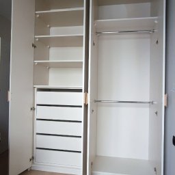 M&R MEBLE - Biała szafa wnękowa z otwartymi drzwiami, widoczne półki, szuflady i drążki na ubrania, na jasnej podłodze.