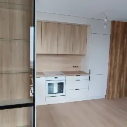 Jasna kuchnia z drewnianymi frontami szafek, białymi blatami i sprzętem AGD, uzupełniona o przeszkloną witrynę z czarną ramą i drewnianymi półkami, na tle jasnej podłogi.