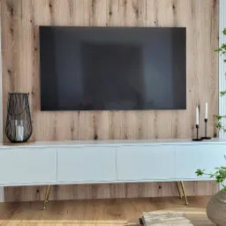 Nowoczesny salon z białą szafką RTV na złotych nóżkach, drewnianą ścianą pod telewizorem i dekoracyjną rośliną w wazonie.