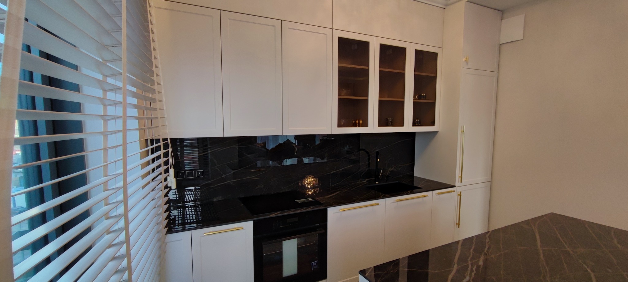 Nowoczesna kuchnia z białymi szafkami, złotymi uchwytami i blatem z czarnego marmuru, oświetlona przez żaluzje okienne.