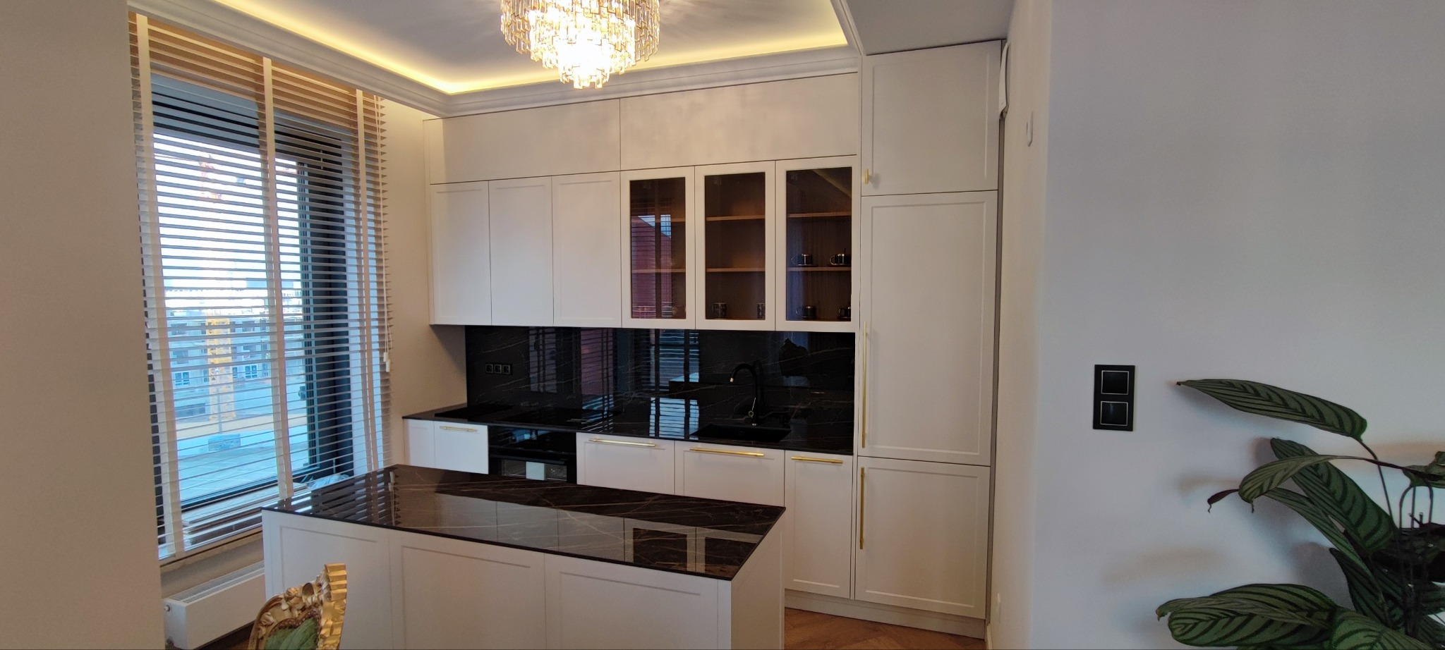 Nowoczesna, jasna kuchnia z wyspą, blatem z czarnego marmuru i złotymi uchwytami na tle okna z żaluzjami.