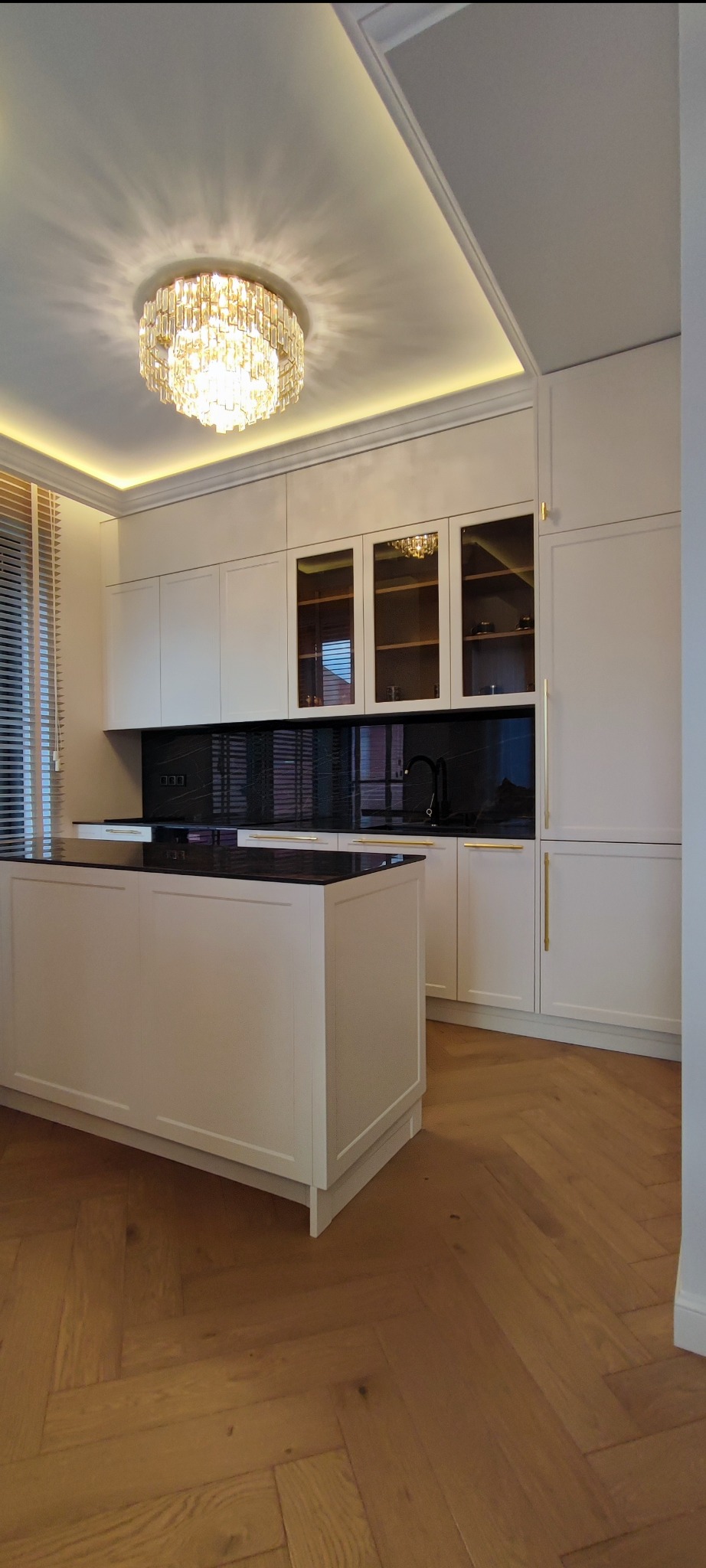 Elegancka kuchnia z białymi szafkami, czarnym blatem i wyspą, oświetlona kryształowym żyrandolem, z parkietem ułożonym w jodełkę.