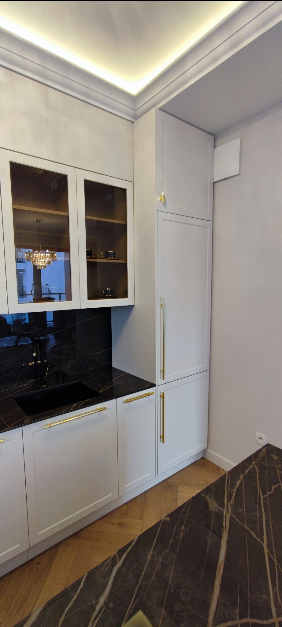 Nowoczesna kuchnia w odcieniach bieli z czarnym blatem imitującym marmur i złotymi uchwytami, widoczne oświetlenie podszafkowe i odbicie żyrandola w szklanych frontach szafek.