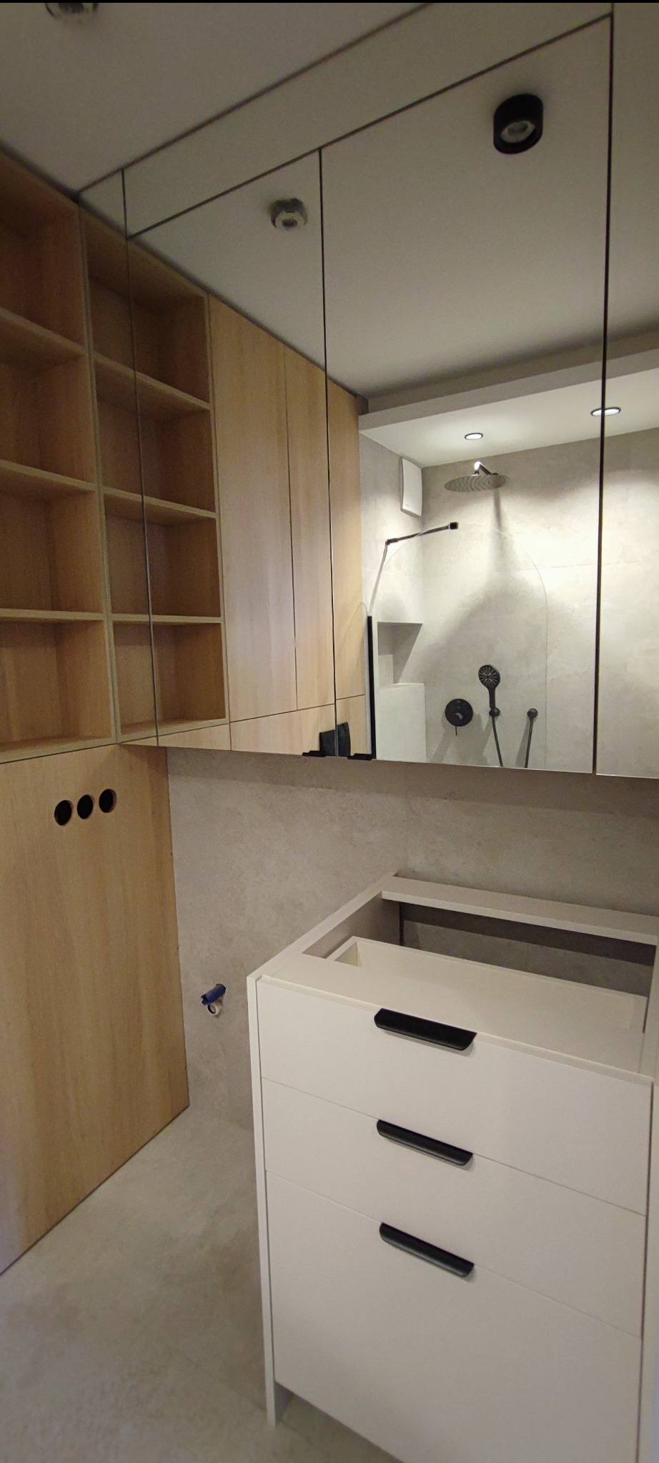 Wnętrze łazienki w trakcie remontu: widoczna szafka pod umywalkę z trzema szufladami, drewniane szafki wiszące z otwartymi półkami i lustro, w którym odbija się kabina prysznicowa z czarną armaturą.