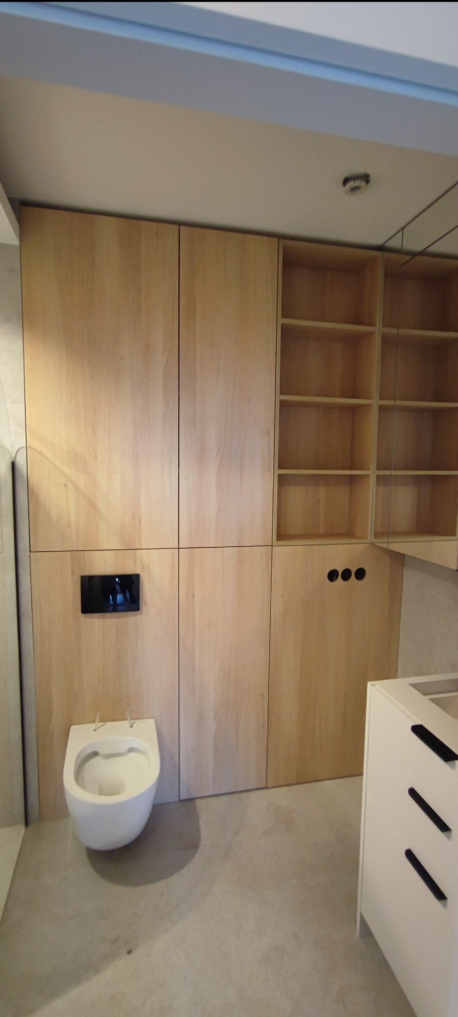 Nowoczesna łazienka z zabudową z jasnego drewna, ukrywającą spłuczkę i tworzącą szafki, z widoczną toaletą podwieszaną i elementami instalacji elektrycznej.