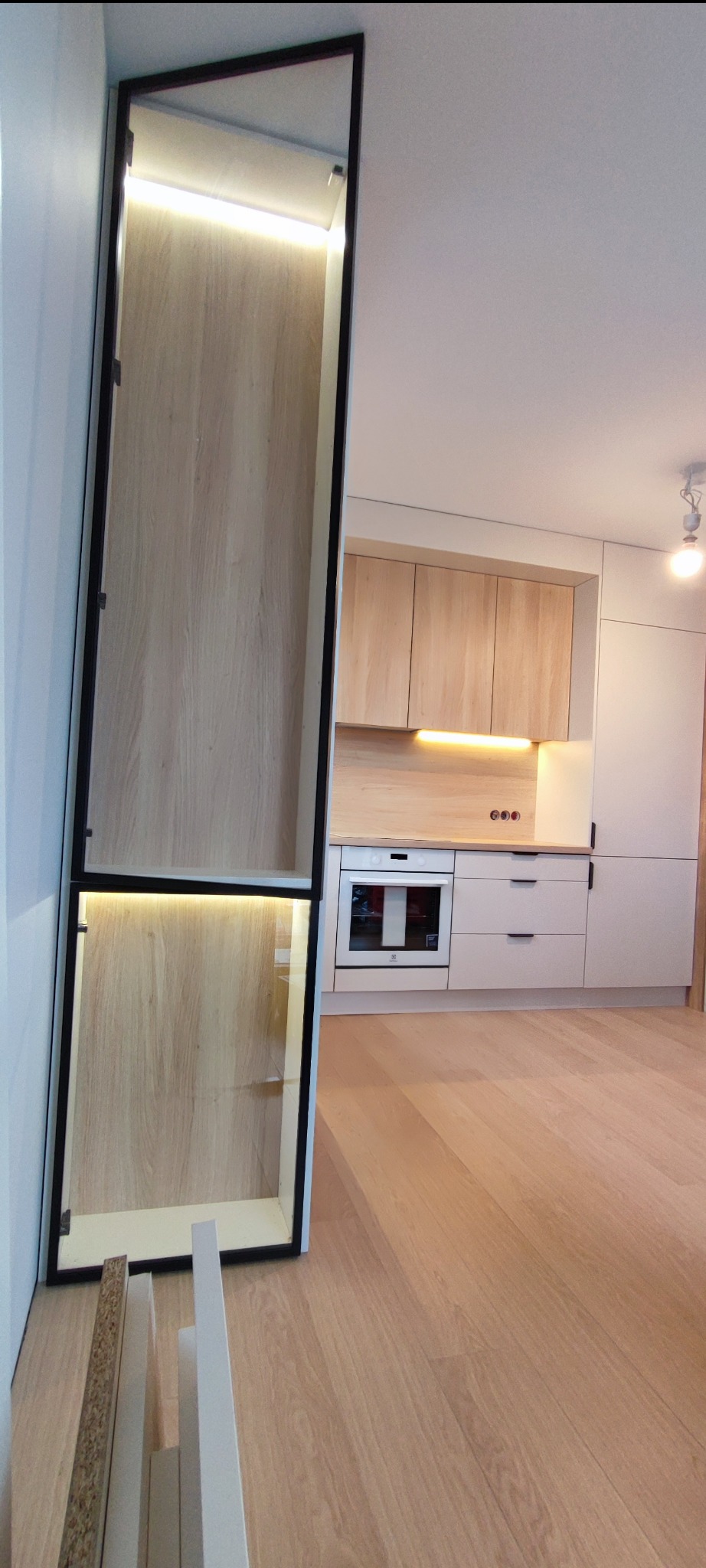 Nowoczesna kuchnia z jasnymi drewnianymi frontami szafek, oświetleniem LED pod szafkami górnymi i wysoką, wąską szafką z czarną ramą i oświetlonymi półkami, z widocznymi elementami montażowymi...