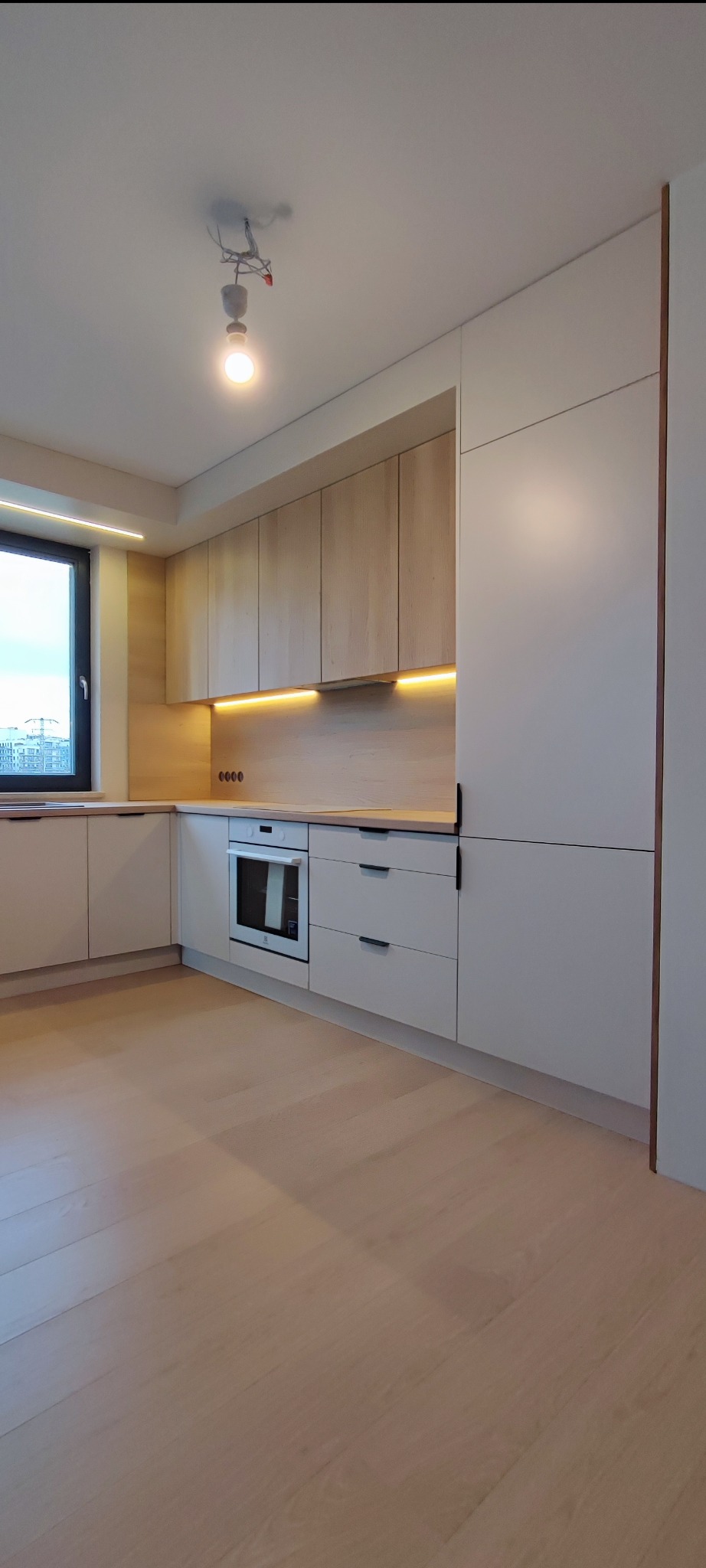 Jasna kuchnia z drewnianymi frontami szafek górnych i białymi dolnymi, oświetlenie LED podszafkowe, minimalistyczne czarne uchwyty, widok na miasto przez okno.