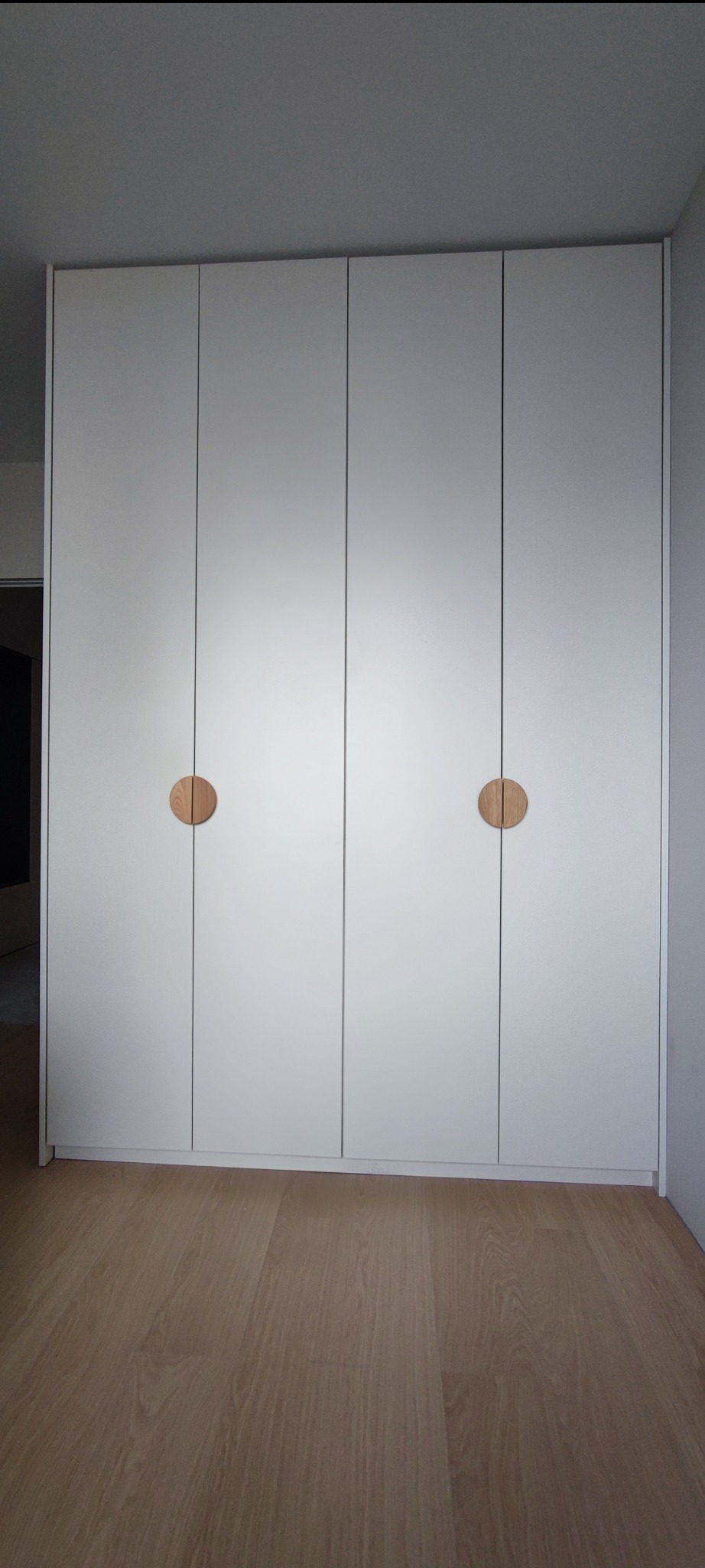 Biała szafa wnękowa z czterema drzwiami i okrągłymi, drewnianymi uchwytami, na drewnianej podłodze.