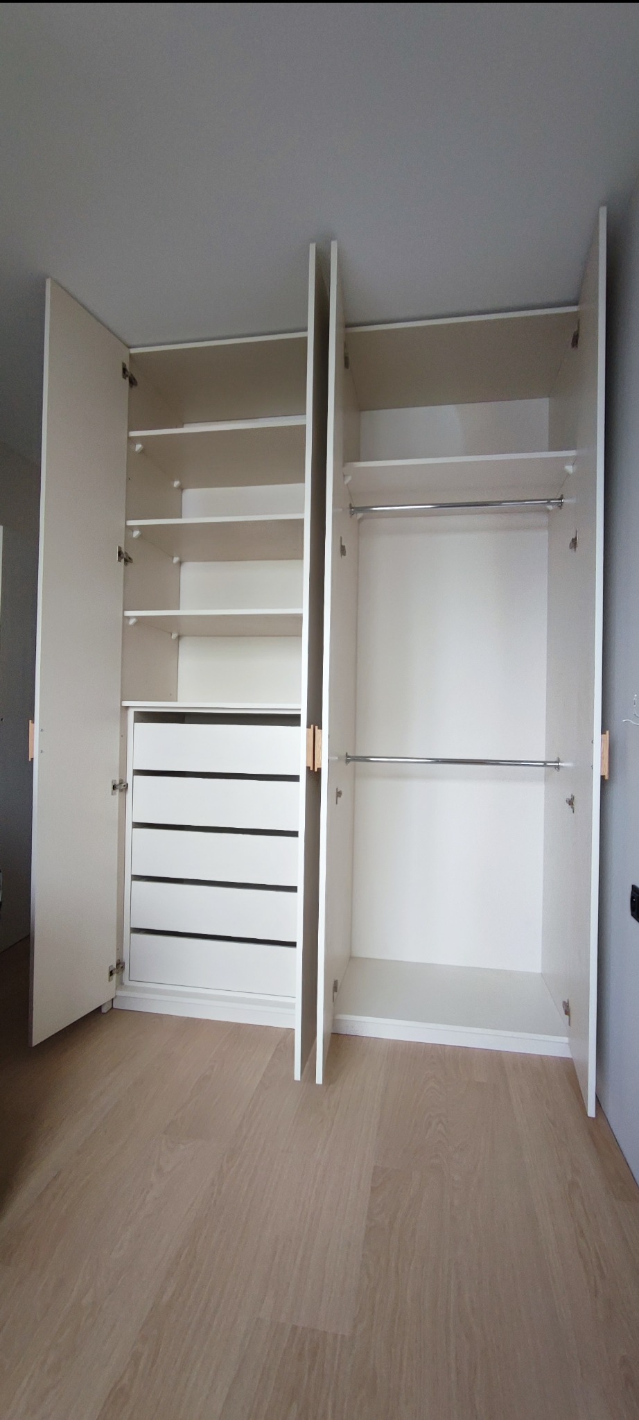 Biała szafa wnękowa z otwartymi drzwiami, widoczne półki, szuflady i drążki na ubrania, na jasnej podłodze.