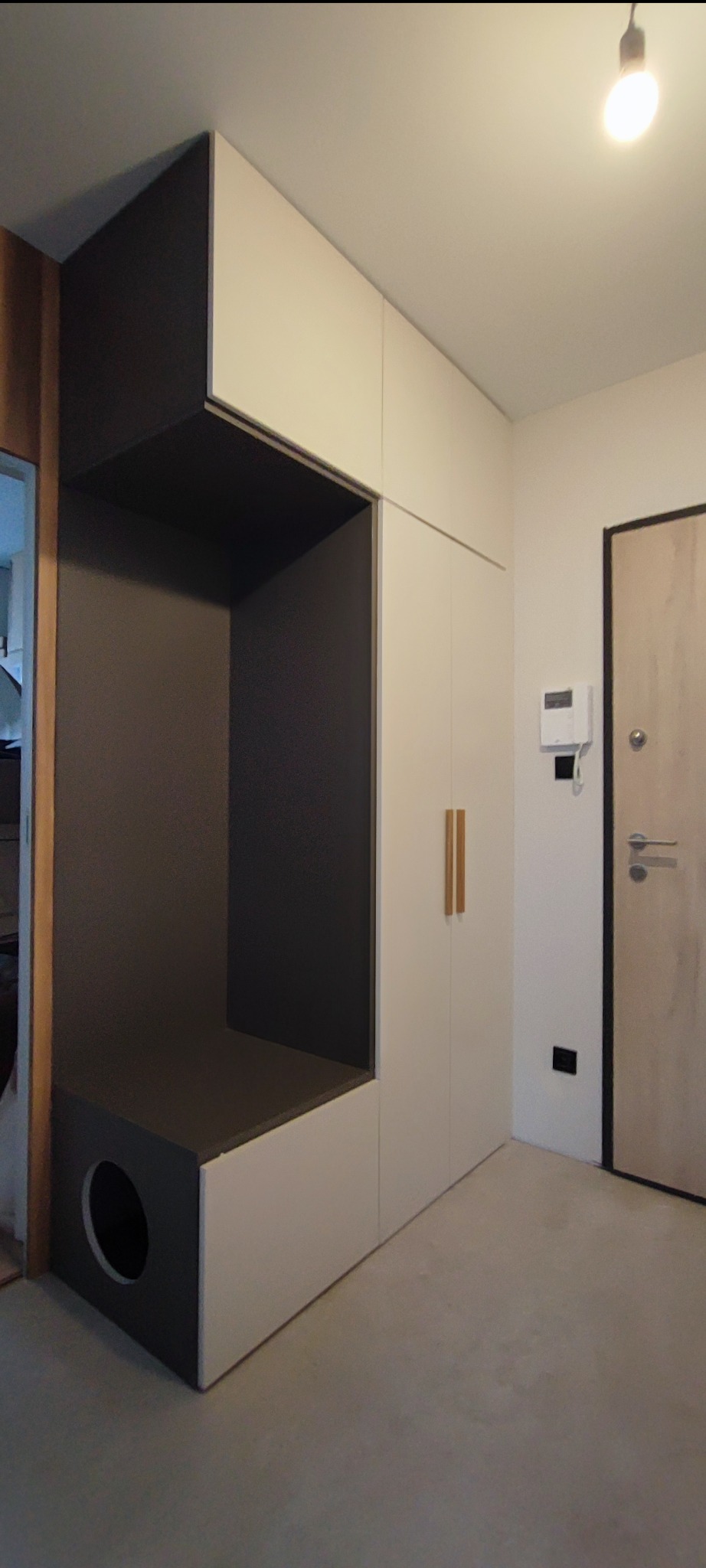 Biało-szara szafa w przedpokoju z wnęką i otworem na legowisko dla zwierząt, minimalistyczne drewniane uchwyty, widok na drzwi wejściowe.