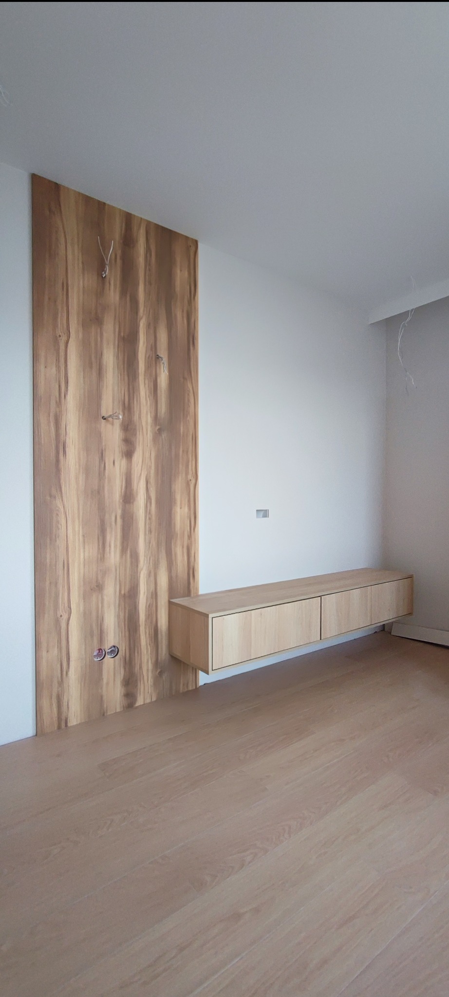 Drewniana ściana z ukrytymi przewodami i wisząca szafka RTV w minimalistycznym wnętrzu z drewnianą podłogą.