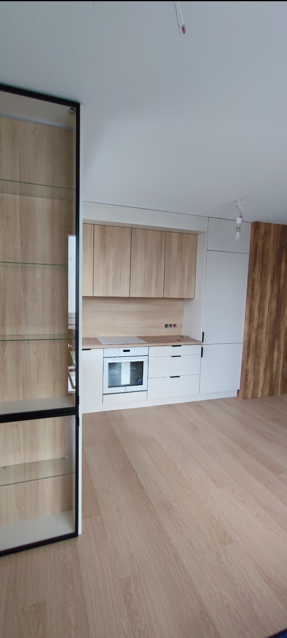 Jasna kuchnia z drewnianymi frontami szafek, białymi blatami i sprzętem AGD, uzupełniona o przeszkloną witrynę z czarną ramą i drewnianymi półkami, na tle jasnej podłogi.