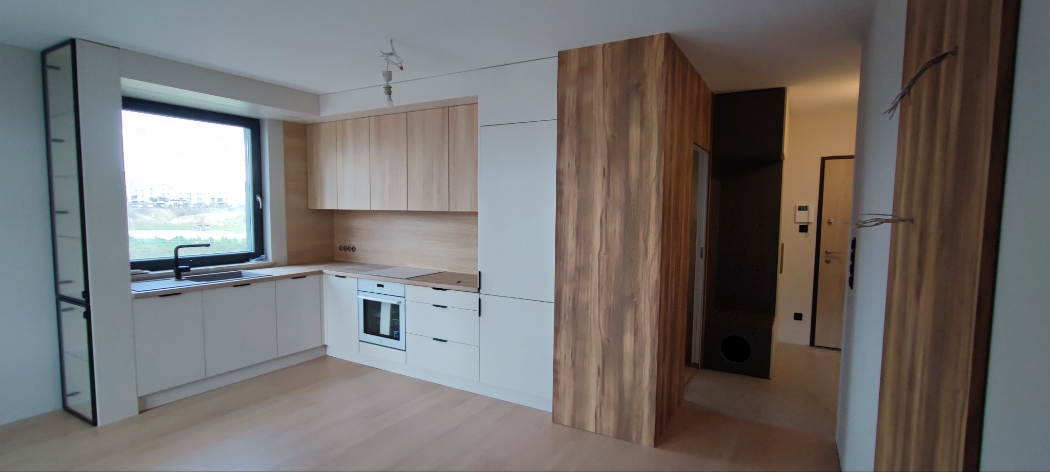 Nowoczesna, jasna kuchnia z białymi frontami i drewnianymi akcentami w Gdańsku. Widoczne okno z czarną ramą, zlewozmywak oraz zabudowana lodówka. Uwagę zwraca drewniana okładzina ściany.