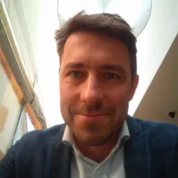 Portret mężczyzny w niebieskiej marynarce i jasnej koszuli, patrzącego w obiektyw, z lampą sufitową w tle.