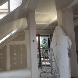 GRZEGORZ Zabagło - Poddasze w trakcie prac wykończeniowych: ściany i skosy pokryte płytami gipsowo-kartonowymi, widoczne okna dachowe i aluminiowa drabina oparta o filar zabezpieczony folią malarską.