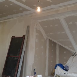 GRZEGORZ Zabagło - Poddasze w trakcie prac wykończeniowych: widoczne płyty karton-gips na skośnym suficie, łączenia zaszpachlowane, przygotowane do dalszych prac. Okno dachowe wpuszcza naturalne światło. W tle...