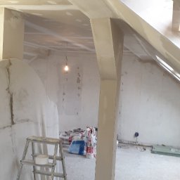 GRZEGORZ Zabagło - Poddasze w trakcie prac wykończeniowych: widoczne elementy konstrukcyjne pokryte gładzią, drabina malarska z taśmą, worki z materiałami budowlanymi i wisząca żarówka.