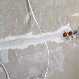 GRZEGORZ Zabagło - Surowa ściana z widocznymi przewodami elektrycznymi i gniazdkami, częściowo pokryta świeżą warstwą wyrównującej zaprawy, przygotowana do dalszych prac wykończeniowych.