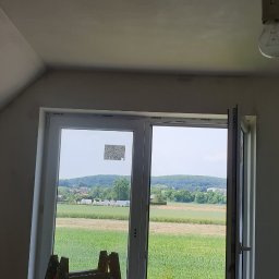 GRZEGORZ Zabagło - Poddasze po nałożeniu gładzi, widoczne ślady po szpachlowaniu na suficie, okno dachowe z widokiem na wieś, żarówka na kablu.