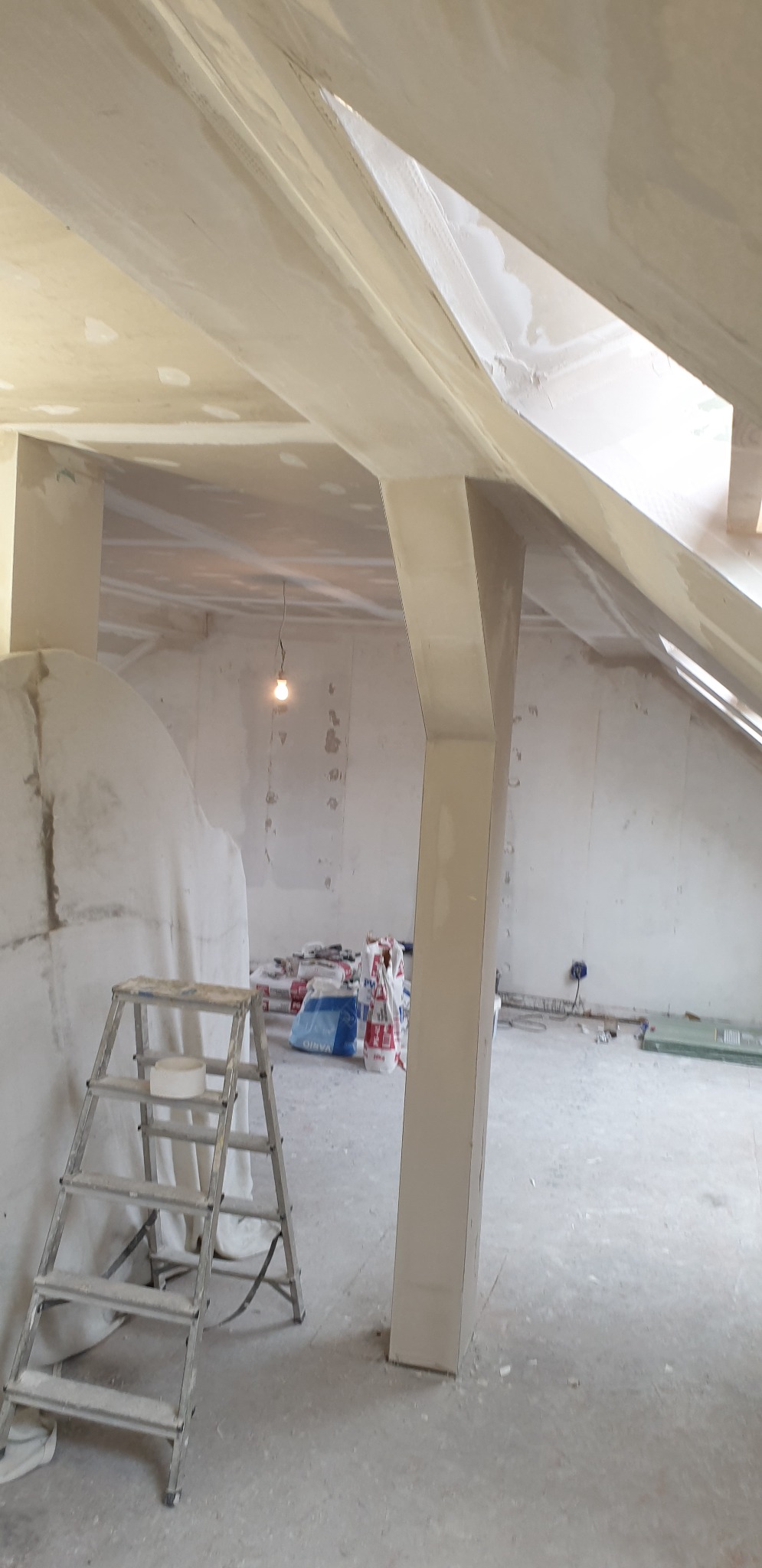 Poddasze w trakcie prac wykończeniowych: widoczne elementy konstrukcyjne pokryte gładzią, drabina malarska z taśmą, worki z materiałami budowlanymi i wisząca żarówka.