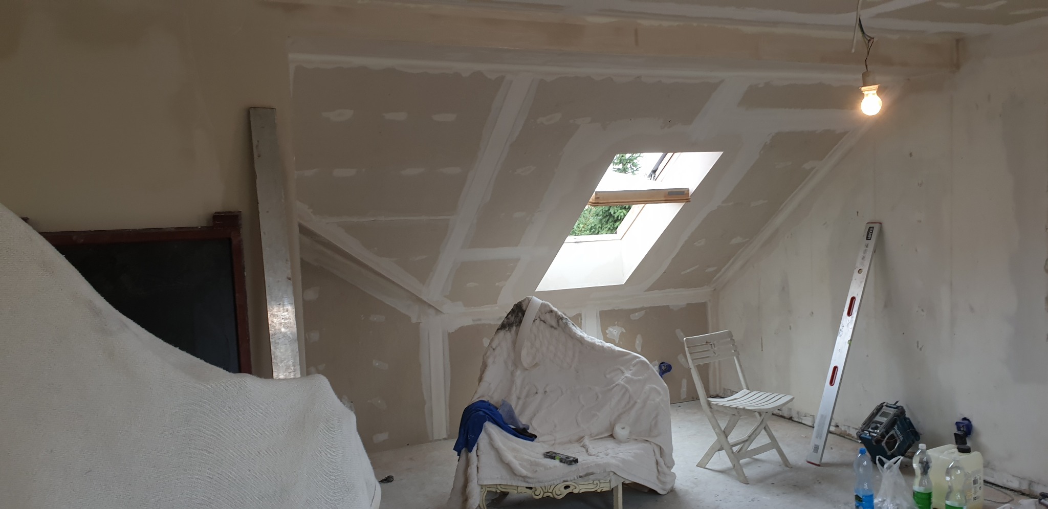 Poddasze w trakcie prac wykończeniowych: widoczne płyty karton-gips na skośnym suficie i ścianach, łączenia szpachlowane, okno dachowe wpuszczające światło, a na podłodze białe krzesło i poziomica...