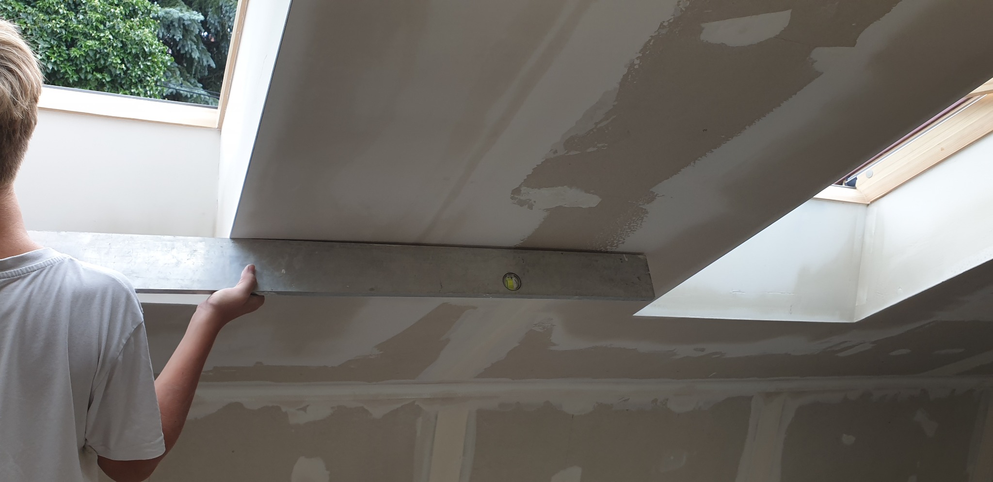 Sprawdzanie poziomu sufitu z płyt kartonowo-gipsowych przy użyciu poziomicy, widoczne łączenia płyt i okno dachowe.