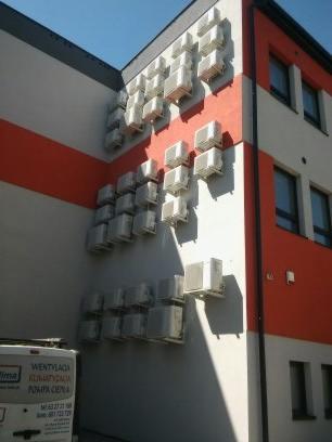 Fasada budynku z licznymi zamontowanymi jednostkami zewnętrznymi klimatyzacji, widoczny fragment samochodu serwisowego z logo firmy.