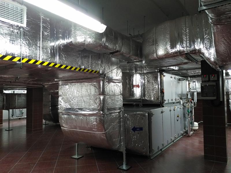 System wentylacyjny z izolacją aluminiową w budynku komercyjnym, widoczne rury i skrzynki wentylacyjne oraz tablica kontrolna na tle brązowych płytek podłogowych i kolumn.
