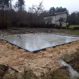 Wylany fundament pod budynek, zabezpieczony czarną folią, otoczony piaskiem, z widocznymi rurami instalacyjnymi i domem w tle.