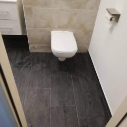 Sebastian Lewczuk - Nowoczesna toaleta wisząca z podtynkową spłuczką, beżowe płytki ścienne i grafitowa podłoga z płytek, uchwyt na papier toaletowy na białej ścianie, widok z poziomu podłogi przez uchylone drzwi.