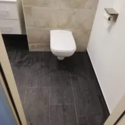 Nowoczesna toaleta wisząca z podtynkową spłuczką, beżowe płytki ścienne i grafitowa podłoga z płytek, uchwyt na papier toaletowy na białej ścianie, widok z poziomu podłogi przez uchylone drzwi.