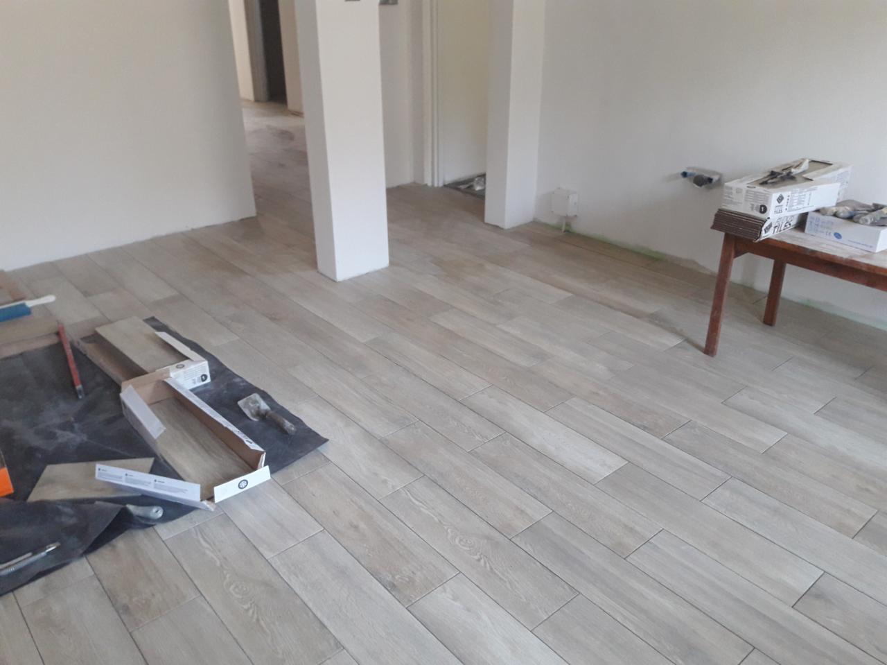 Układanie jasnych płytek podłogowych imitujących drewno w nowym pomieszczeniu, widoczne narzędzia i kartony płytek oraz białe filary.