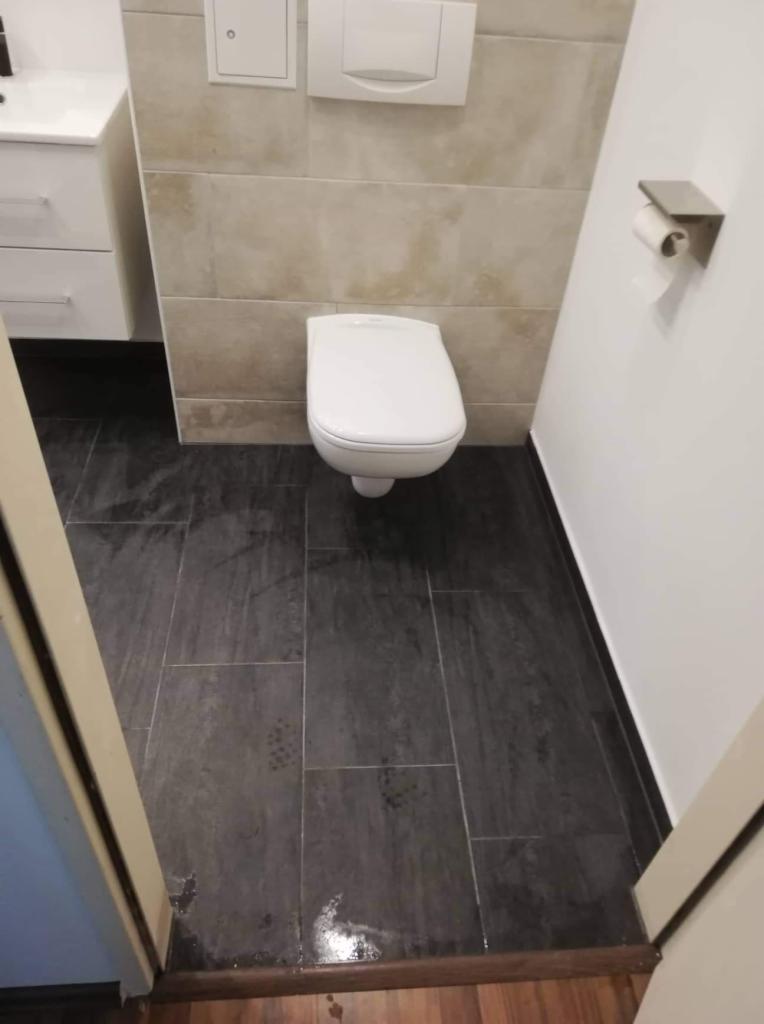 Nowoczesna toaleta wisząca z podtynkową spłuczką, beżowe płytki ścienne i grafitowa podłoga z płytek, uchwyt na papier toaletowy na białej ścianie, widok z poziomu podłogi przez uchylone drzwi.