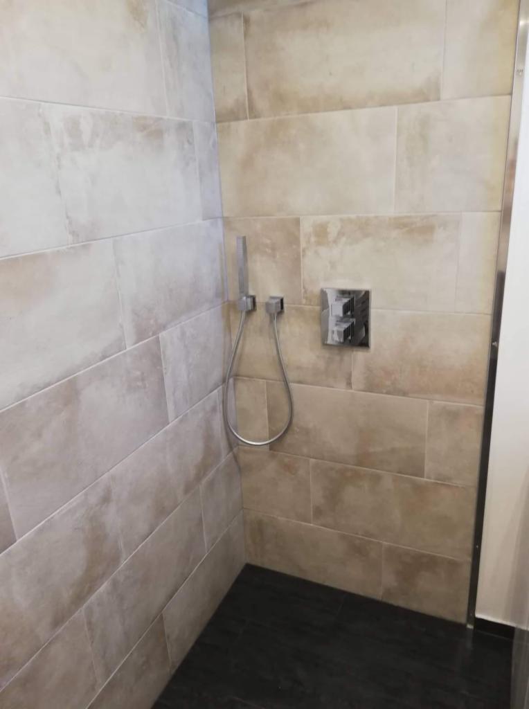 Kabina prysznicowa z beżowymi, prostokątnymi płytkami imitującymi kamień, ciemną podłogą i chromowaną armaturą.