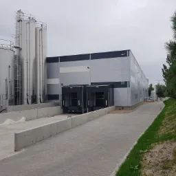 Nowoczesna hala przemysłowa z rampami załadunkowymi i stalowymi silosami, widoczni pracownicy w uniformach przy silosach.