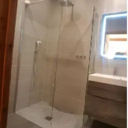 Nowoczesna kabina prysznicowa z deszczownicą i szklaną ścianką, umywalka z szafką w kolorze drewna, podświetlane lustro, jasne płytki na ścianach i mozaika na podłodze prysznica.