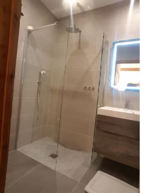 Nowoczesna kabina prysznicowa z deszczownicą i szklaną ścianką, umywalka z szafką w kolorze drewna, podświetlane lustro, jasne płytki na ścianach i mozaika na podłodze prysznica.