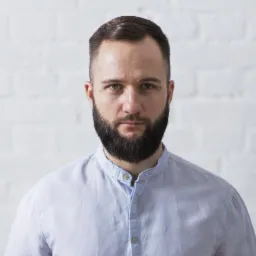 Portret brodatego mężczyzny w błękitnej koszuli na tle białej ściany z cegieł.