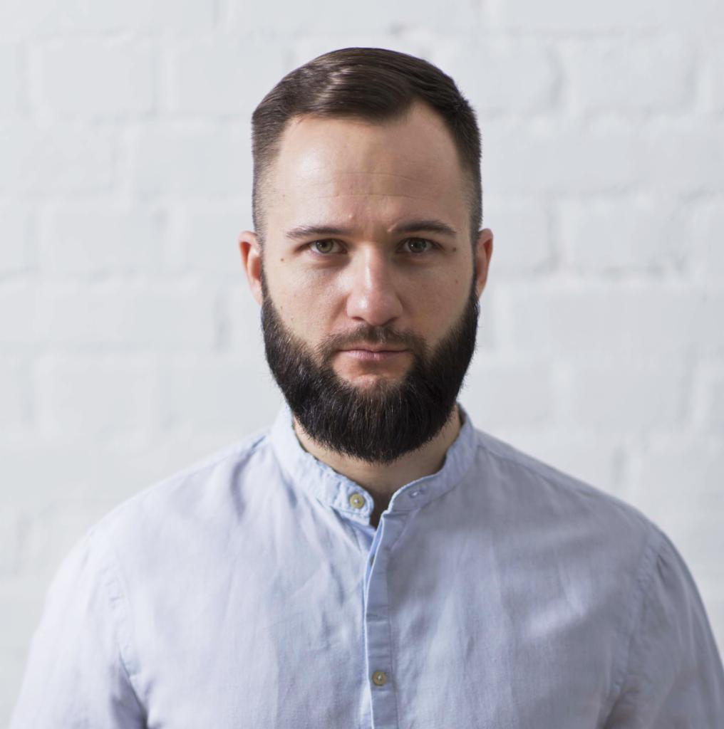 Portret brodatego mężczyzny w błękitnej koszuli na tle białej ściany z cegieł.