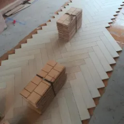 Układanie jasnych paneli podłogowych w jodełkę, widoczne stosy paneli gotowych do montażu i fragment podłoża z klejem.