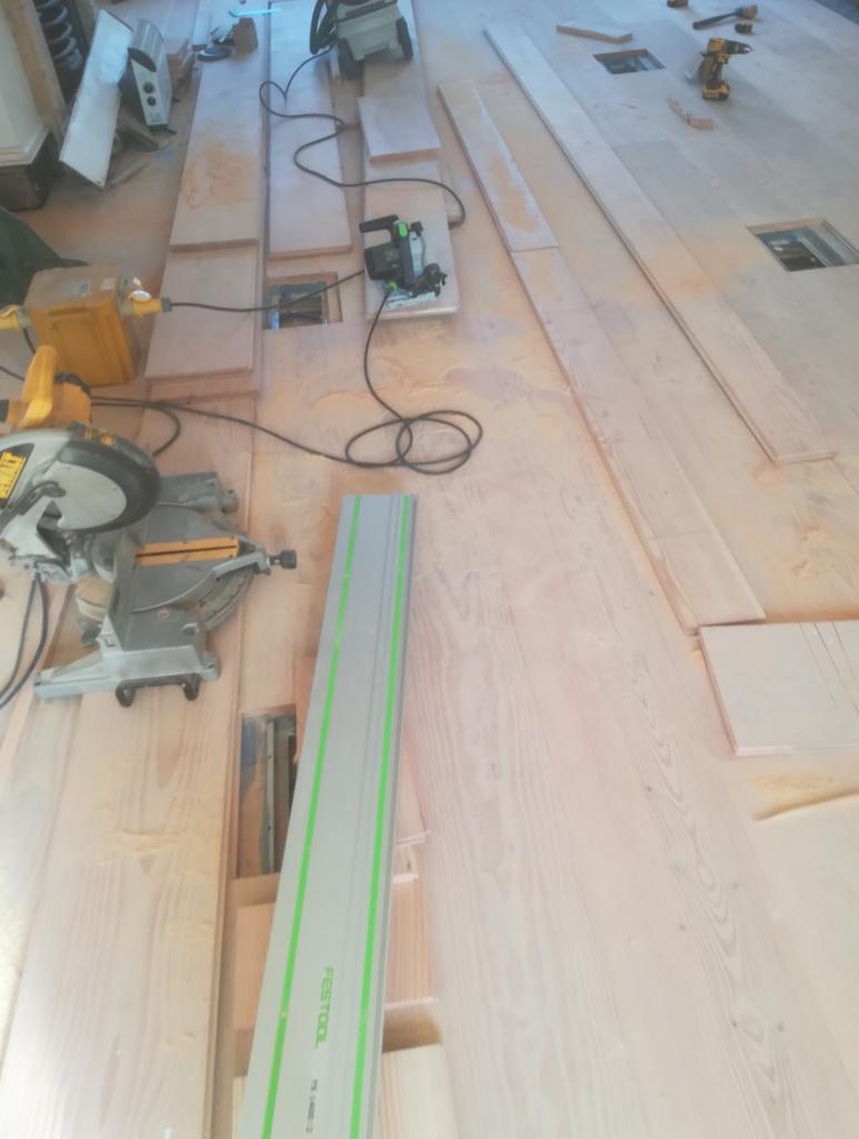 Przygotowanie do ułożenia jasnych paneli podłogowych, widoczne narzędzia: piła ukośnica, szyna prowadząca, wyrzynarka i wiertarka akumulatorowa, fragment podłogi z otworami wentylacyjnymi.