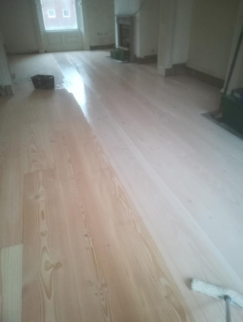 Świeżo polakierowana, jasna podłoga drewniana z widocznym wałkiem malarskim w rogu pomieszczenia, odbijająca światło z okna.