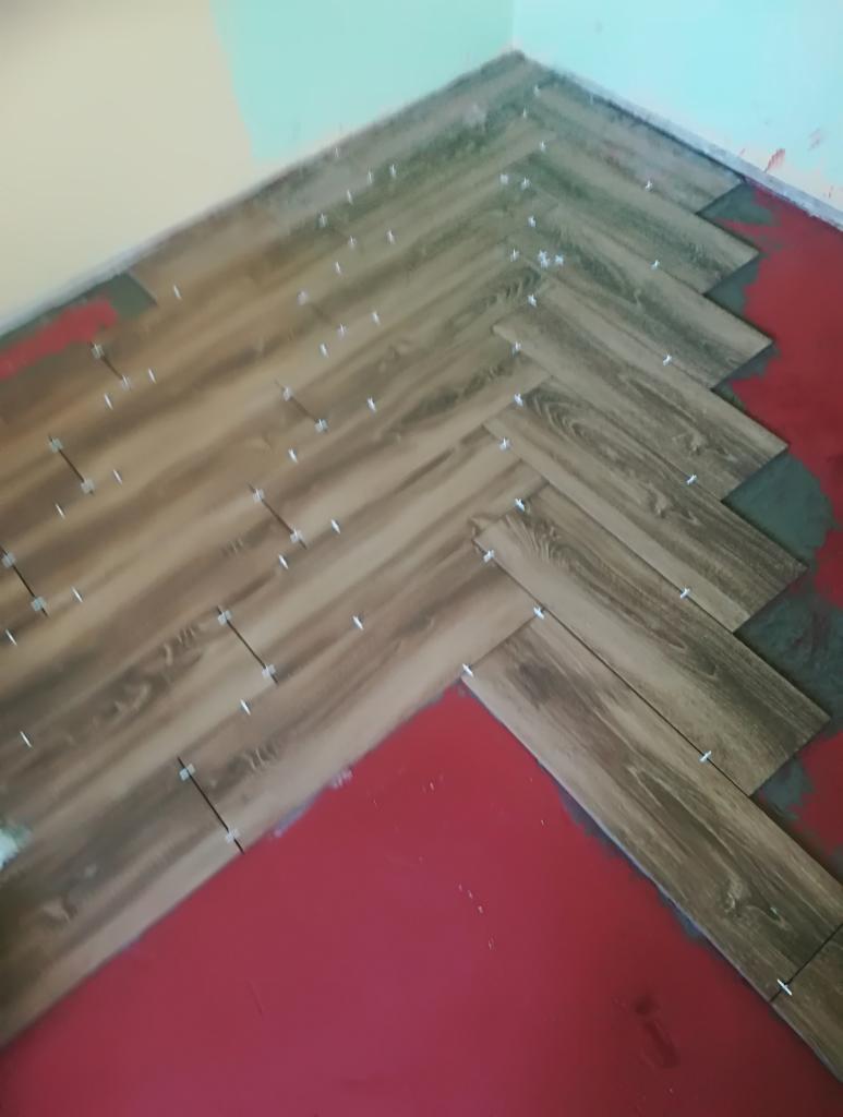 Układanie paneli podłogowych w jodełkę, widoczne plastikowe krzyżyki dystansowe i czerwony podkład pod panele.