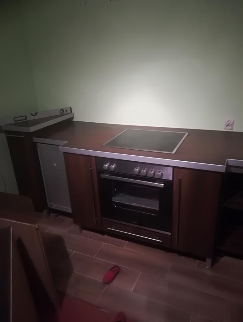 Ciemna kuchnia z brązowymi szafkami, piekarnikiem i płytą grzewczą, widoczne niedopasowane panele podłogowe i czerwone kapcie.